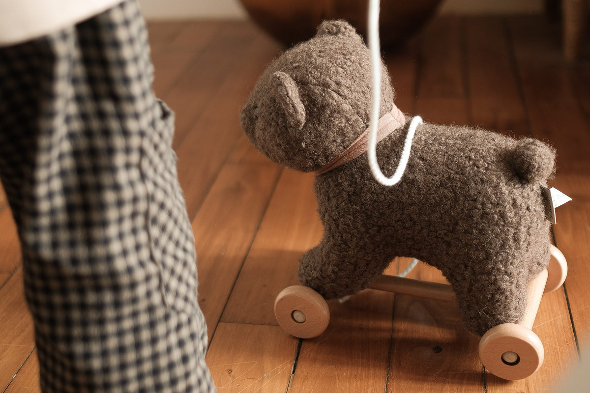 A child pulls Raoul, the plush pull-along bear on wooden wheels, across a wooden floor