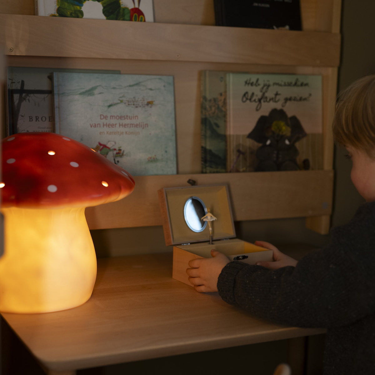 A child opens a musical box at a desk, next to a large red mushroom lamp that is switched on
